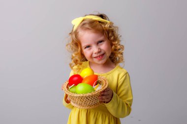 Küçük kıvırcık saçlı bir kız, gri arka planda Paskalya yumurtaları olan sepeti tutuyor.