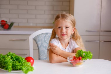 Sevimli sarışın çocuk mutfakta oturmuş sebze yiyor.
