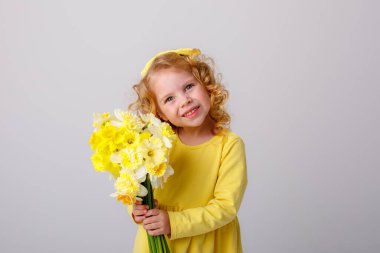 Sarı elbiseli, küçük kıvırcık saçlı bir kız elinde bir buket bahar çiçeği tutuyor.  