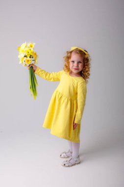 Sarı elbiseli, küçük kıvırcık saçlı bir kız elinde bir buket bahar çiçeği tutuyor.  