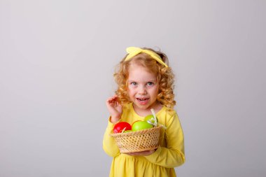 Küçük kıvırcık saçlı bir kız, gri arka planda Paskalya yumurtaları olan sepeti tutuyor.