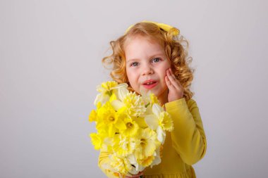 Sarı elbiseli, küçük kıvırcık saçlı bir kız elinde bir buket bahar çiçeği tutuyor.  