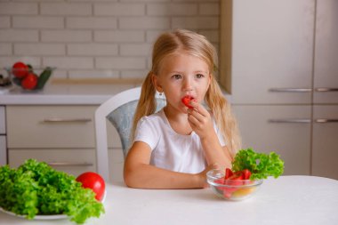 Sevimli sarışın çocuk mutfakta oturmuş sebze yiyor.