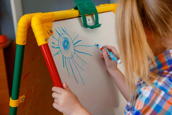 smiling child girl blonde draws on a white Board with a felt-tip pen at home