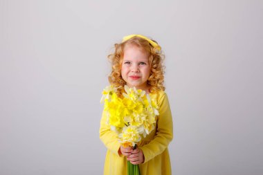 Sarı elbiseli, küçük kıvırcık saçlı bir kız elinde bir buket bahar çiçeği tutuyor.  