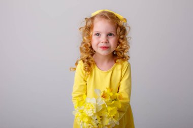 Sarı elbiseli küçük kıvırcık saçlı bir kız elinde gri arka planda bir buket bahar çiçeği tutuyor. 