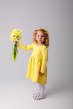 Sarı elbiseli, küçük kıvırcık saçlı bir kız elinde bir buket bahar çiçeği tutuyor.  