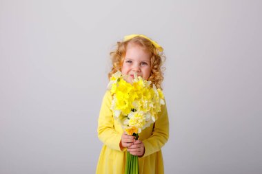 Sarı elbiseli küçük kıvırcık saçlı bir kız elinde gri arka planda bir buket bahar çiçeği tutuyor. 