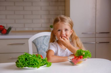 Sevimli sarışın çocuk mutfakta oturmuş sebze yiyor.