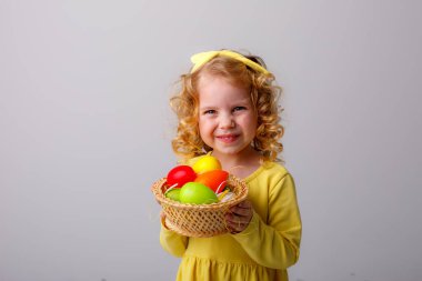 Küçük kıvırcık saçlı bir kız, gri arka planda Paskalya yumurtaları olan sepeti tutuyor.