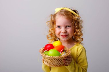 Küçük kıvırcık saçlı bir kız, gri arka planda Paskalya yumurtaları olan sepeti tutuyor.