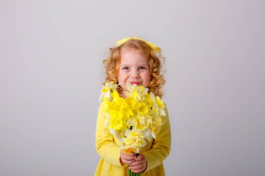 Sarı elbiseli, küçük kıvırcık saçlı bir kız elinde bir buket bahar çiçeği tutuyor.  