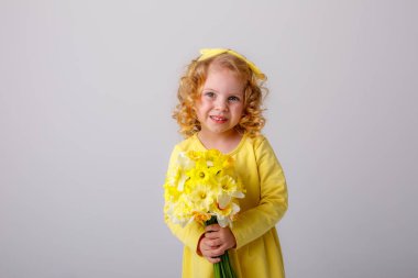 Sarı elbiseli küçük kıvırcık saçlı bir kız elinde gri arka planda bir buket bahar çiçeği tutuyor. 