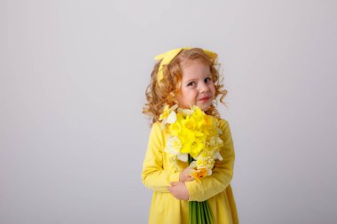 Sarı elbiseli, küçük kıvırcık saçlı bir kız elinde bir buket bahar çiçeği tutuyor.  