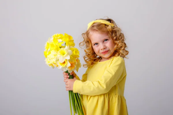 一个卷曲卷曲的小女孩 穿着一件黄色的衣服 手里拿着一束灰色背景的春花 — 图库照片