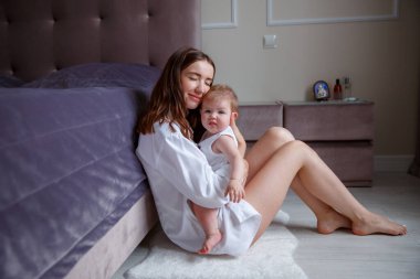 Güzel, genç bir anne kucağında bebeği, evde yatağın yanında oturuyor. 