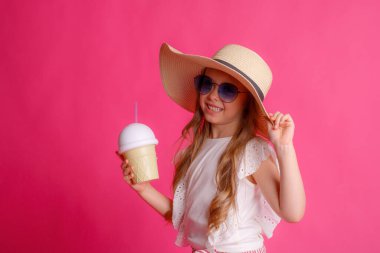 Güneş gözlüklü küçük bir sarışının portresi pembe arka planda bir şapka ve soğutucu bir kokteyl kutusu.