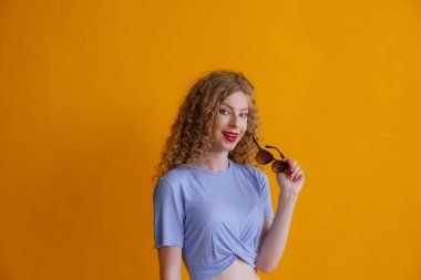 Turuncu arka planda güneş gözlüklü kıvırcık saçlı kadın poz veriyor.