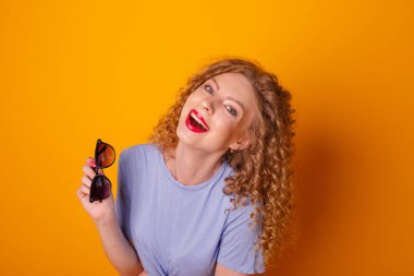 Turuncu arka planda güneş gözlüklü kıvırcık saçlı kadın poz veriyor.