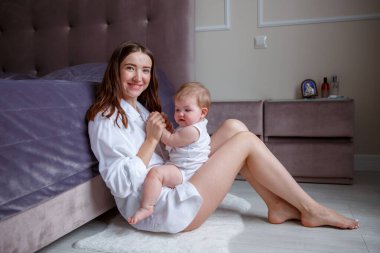 Güzel, genç bir anne kucağında bebeği, evde yatağın yanında oturuyor. 