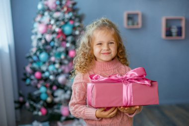 Noel ağacının yanındaki küçük kız elinde gülümseyerek bir hediye tutuyor. 