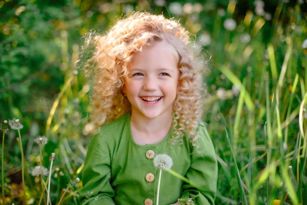 小さな巻き毛のブロンド女の子で緑のドレス吹いてタンポポで公園の笑顔 — ストック写真