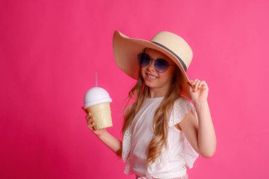 Güneş gözlüklü küçük bir sarışının portresi pembe arka planda bir şapka ve soğutucu bir kokteyl kutusu..
