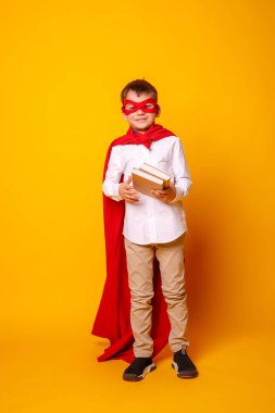 Kırmızı pelerinli çocuk, elinde turuncuya karşı poz veren kitaplar var.