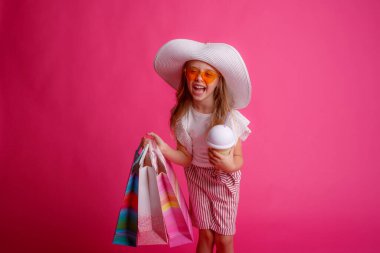Bir sürü alışveriş torbası ve içkiyle küçük bir kız stüdyoda pembe bir arka planda poz veriyor. 