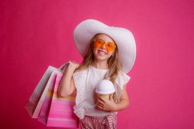 Bir sürü alışveriş torbası ve içkiyle küçük bir kız stüdyoda pembe bir arka planda poz veriyor. 