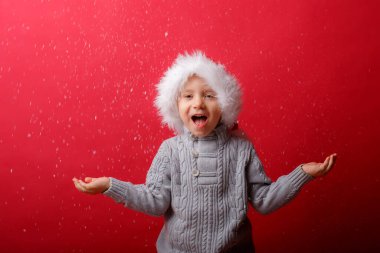 Noel Baba şapkalı bir çocuk kırmızı arka planda kar yakalıyor.
