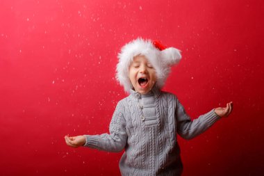 Noel Baba şapkalı bir çocuk kırmızı arka planda kar yakalıyor.