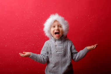 Noel Baba şapkalı bir çocuk kırmızı arka planda kar yakalıyor.