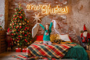 Küçük Asyalı kız, yatak odasında pijamalarıyla, minderlerle, Noel 'le, yılbaşıyla oynaşıyor.