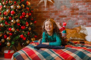 Yatak odasında pijamalı küçük Asyalı kız Noel ağacının yanında, Noel ağacının yanında yatıyordu.