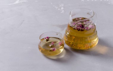 Cam bir çaydanlık ve gül yapraklarından yapılmış demlenmiş küçük bir bardak gri beton arka planda duruyor, seçici bir odak noktası var.