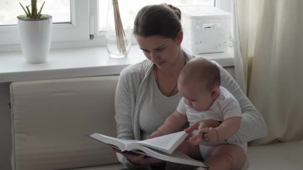 Medycyna, opieka zdrowotna, rodzina, niewinność, pediatria, szczęście, koncepcje niemowląt - młoda szczęśliwa matka czytać książki i bawić się z pulchny 7 miesięcy dziecko siedzi w jasnym słońcu żłobek na miękkim białym łóżku — Wideo stockowe