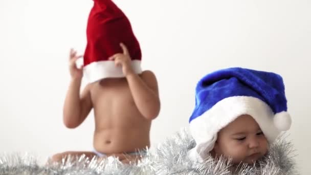 Καλά Χριστούγεννα και ευτυχισμένο το νέο έτος, βρέφος, παιδική ηλικία, έννοια των διακοπών - close-up 6 μηνών νεογέννητο γυμνό μωρό σε κόκκινο και μπλε καπέλο Σάντα Claus με μεγαλύτερη αδελφή σέρνεται με διακοσμήσεις χριστουγεννιάτικο δέντρο — Αρχείο Βίντεο