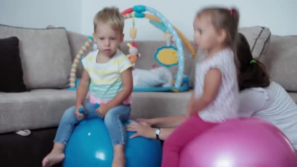Childhood, games, upbringing and development concept - general plan of little children boy brother and girl sister with mom jumping on fitball in kid playroom. Siblings have fun tat home on quarantine — Stock Video