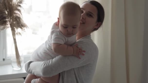 Medicin, sjukvård, familj, oskuld, pediatrik, lycka, spädbarnskoncept - Ung lycklig mor höjer och leker med 7 månaders baby kyssar, smekningar sitter i starkt solljus plantskola nära fönstret — Stockvideo