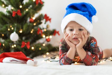 Mutlu Noeller ve mutlu yıllar, çocukluk, tatil konsepti - Göbek oyununda mavi Noel Baba şapkalı küçük üzgün kız ve Noel ağacı süsleriyle yatakta iyi eğlenceler..