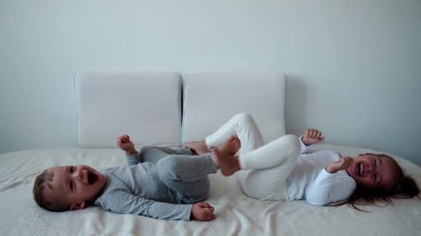 Rodina, nevinnost, kojenecké představy - Dvě usměvavé děti cvičí nohy ležící na bílé posteli. Sourozenci malý chlapec a dívka bratr a sestra se baví a smějí, šťastné děti v karanténě doma. — Stock video