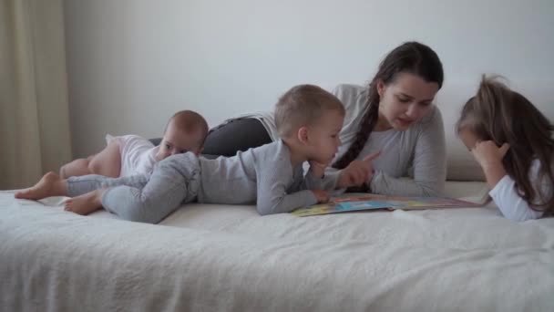 Παιδική ηλικία, μητρότητα, αδέλφια έννοια - Carefree νεαρή μαμά διαβάσει βιβλίο νεράιδα ουρές για τα μικρά παιδιά, ο γιος νεογέννητο μωρό διασκεδάσουν παίζουν στο κρεβάτι, χαρούμενος χαρούμενη οικογένεια γέλιο γαργαλάει, φιλί, αγκαλιές ξαπλωμένος χαλαρώστε — Αρχείο Βίντεο