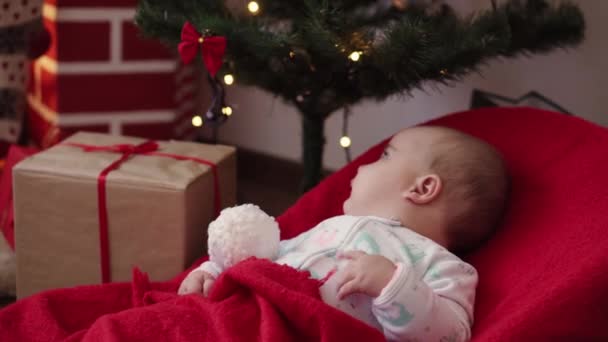 Vánoce, zima, Nový rok, Oslava, rodinný koncept - Krásné usměvavé miminko oblečené do vánočního oblečení sedící v houpacím křesle zábaly v teplé měkké červené přikrývce u krbu a vánočního stromečku. — Stock video