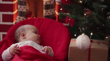 Xmas, kış, yeni yıl, kutlama, aile konsepti Noel kıyafetleri içinde sallanan sandalyeye oturmuş şöminenin ve Noel ağacının yanında sıcak kırmızı battaniyeye sarılmış güzel bir bebek..