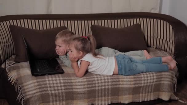 Семья, игры, карантин, детские концепции - двое счастливых маленьких детей смотрят мультики на ноутбуке, лежащем на коричневом диване дома во время карантина. Дети брат и сестра весело провести время вместе — стоковое видео