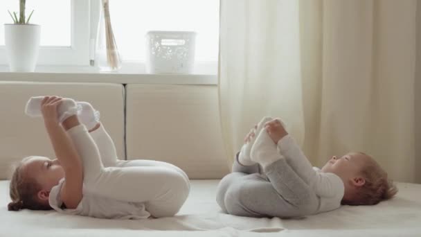 가족, 무죄, 유아 개념 - 두 명의 미소짓는 어린이가 흰 침대에 누워 다리 운동을 한다. 십 대 소년, 소녀, 자매재밌게 웃고 행복 한 아이들 집에 격리 격리. — 비디오