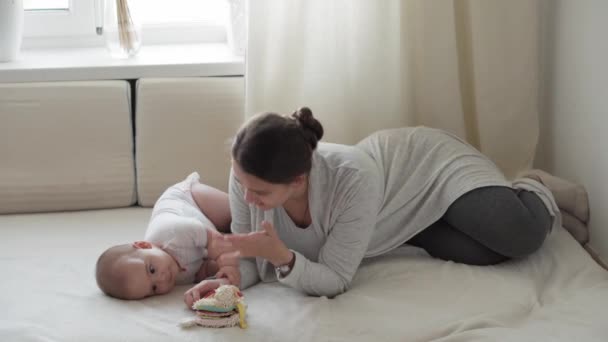 Medicina, salud, familia, inocencia, pediatría, felicidad, conceptos infantiles - Joven madre feliz leer el libro y jugar con gordito bebé de 7 meses sentado en la luz del sol brillante guardería en la cama blanca suave. — Vídeos de Stock