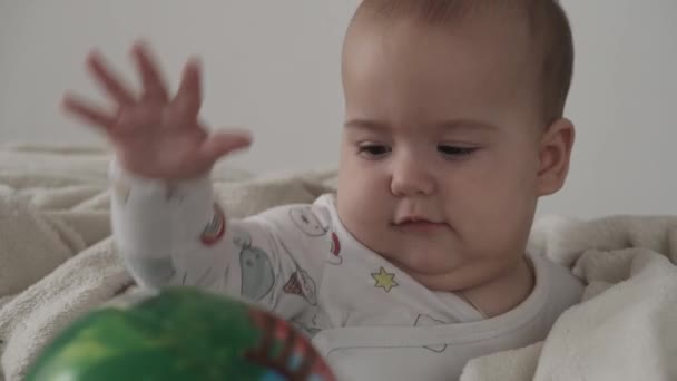 Kojenecká, dětská představa - detailní záběr roztomilého usměvavého veselého hnědookého baculatého novorozence vzhůru. bezzubé 7 měsíců baby play s míčkem sedí na bílé měkké posteli zabalené v teplé dece doma — Stock video