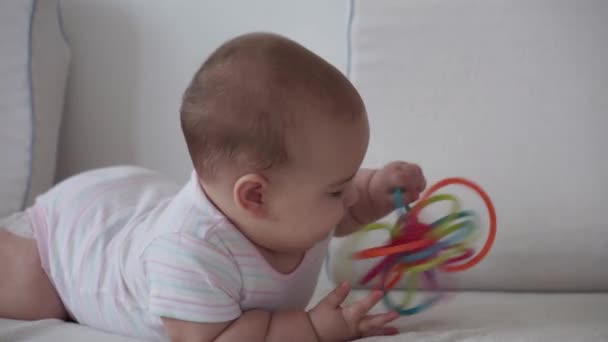 Medycyna, opieka zdrowotna, rodzina, niewinność, pediatria, koncepcje niemowląt - zabawna szczęśliwa 7-miesięczna dziewczynka roi się od zabawek ząbkujących na brzuchu i kołyszących się na kolanach w jasnym słonecznym żłobku na białym miękkim łóżku — Wideo stockowe